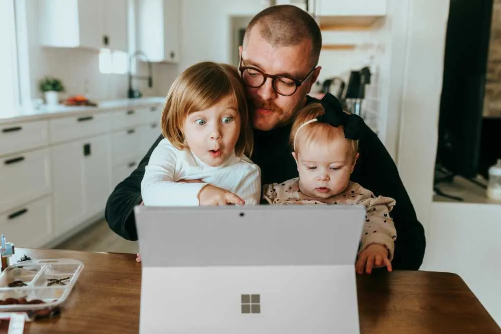 A father holding two young children while they look at a Surface laptop on a kitchen table