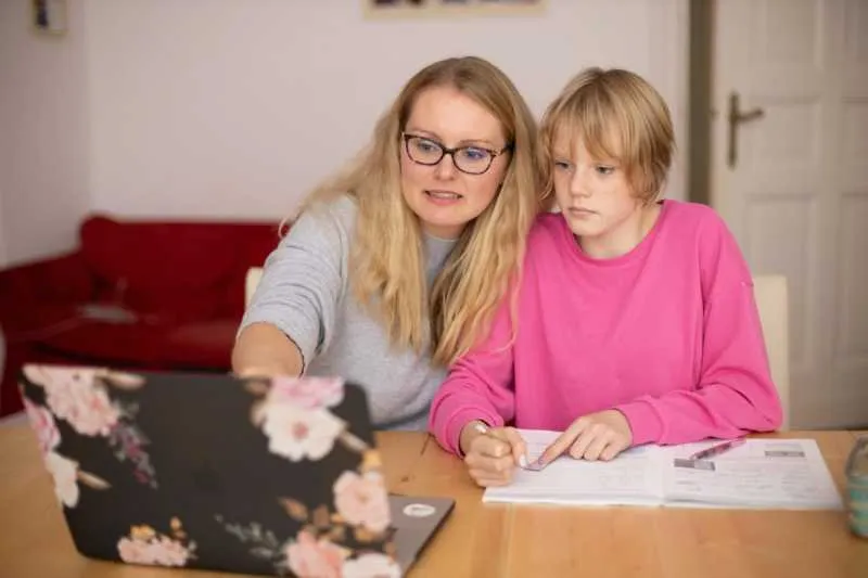 A parent helping a child with schoolwork at a dining table beside an open laptop
